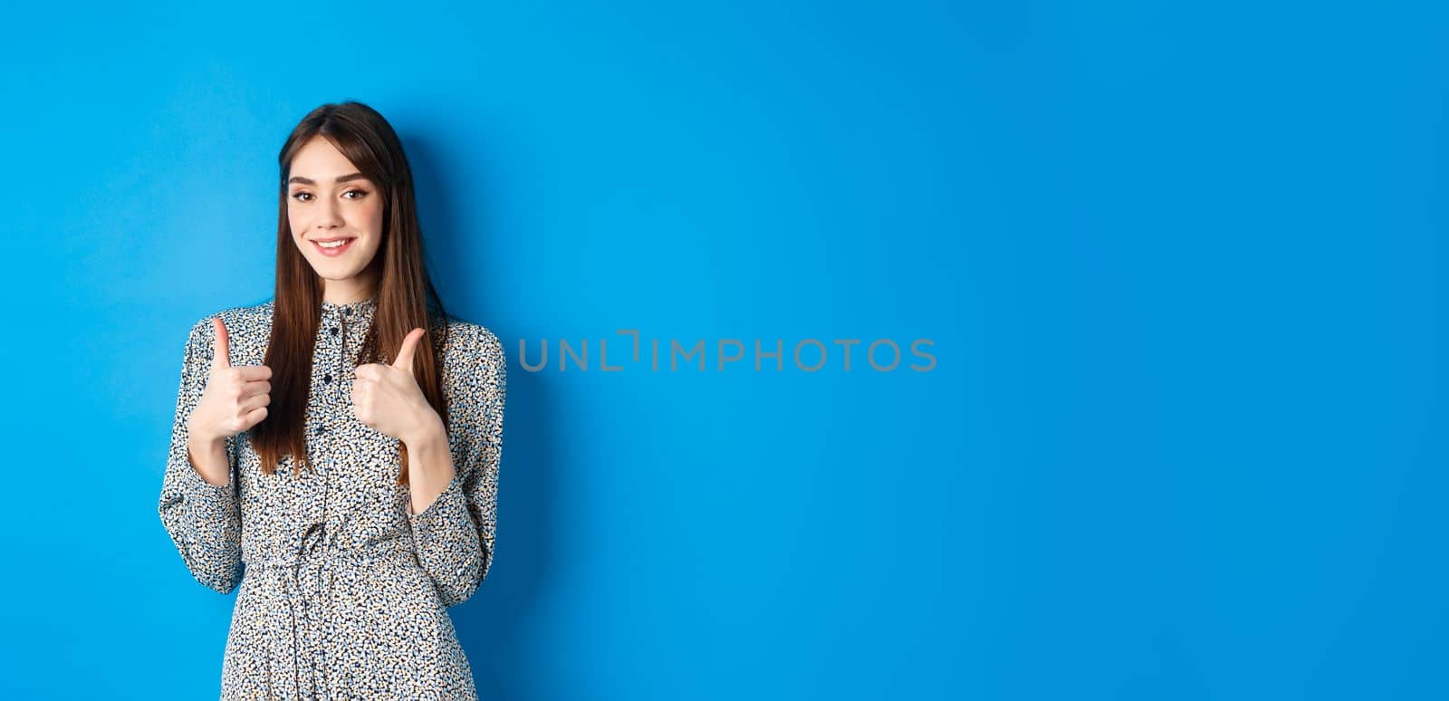 Cheerful smiling woman in dress, showing thumb up and look satisfied, approve and like good thing, recommending product, standing on blue background.