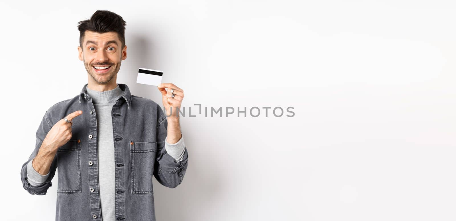 Excited handsome guy with moustache pointing finger at plastic credit card, smiling pleased, recommend good deal, standing on white background.