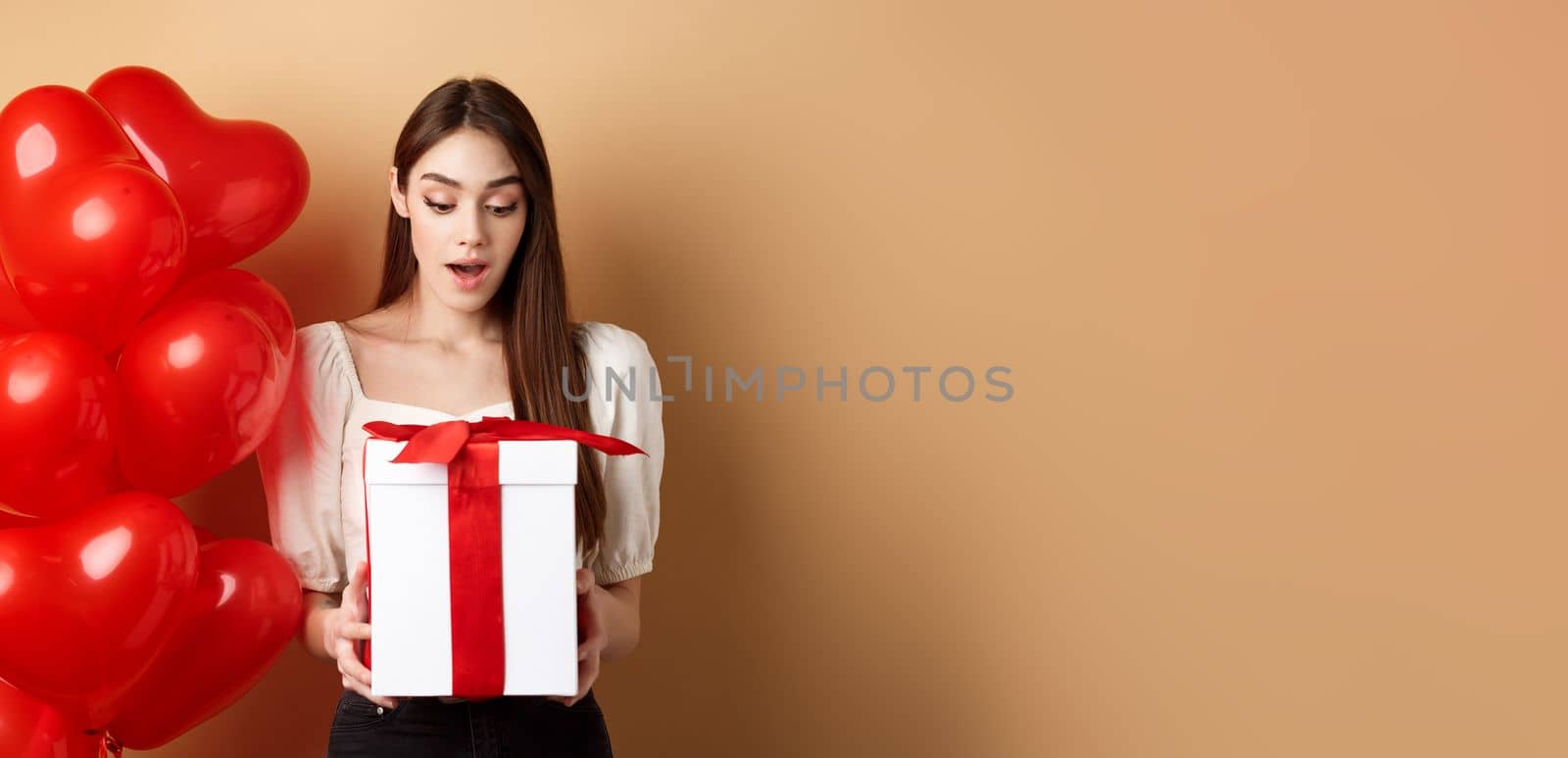 Surprised beautiful woman in romantic outfit, standing near heart balloons and looking at her gift on Valentines day, standing on beige background.