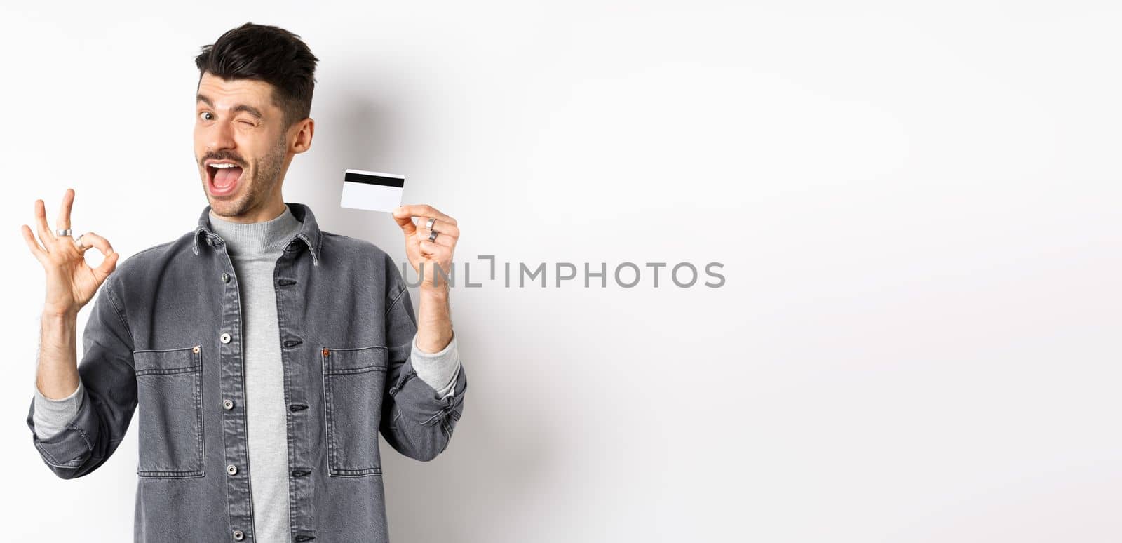 Very good. Smiling guy with plastic credit card showing okay sign, smiling and winking satisfied, recommend bank, standing on white background.