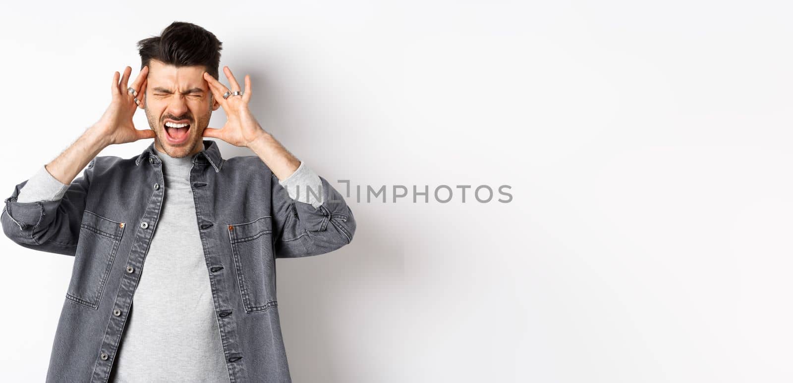 Frustrated young man shouting and touching head distressed, standing bothered by loud noise or headache, standing on white background.