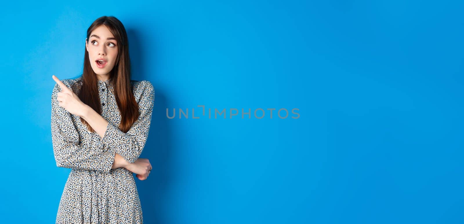 Wow look there. Excited pretty lady in dress looking and pointing left wondered, checking out advertisement, standing on blue background by Benzoix