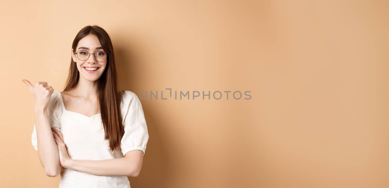 Cheerful woman in eyewear pointing finger left at logo, smiling happy, standing on beige background.