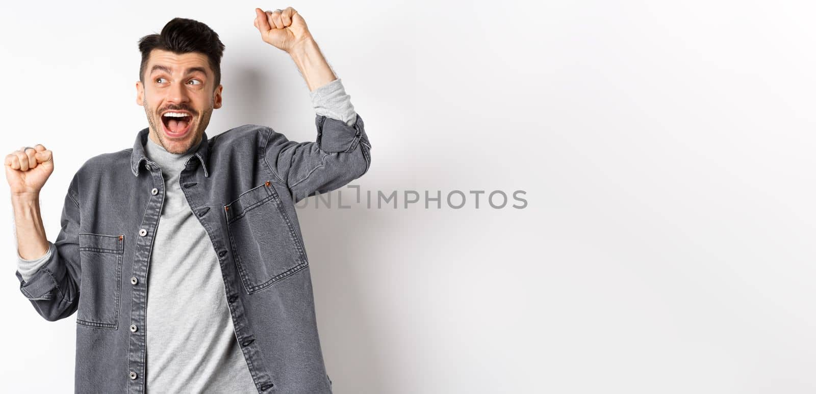 Excited young man cheering and looking aside, winning prize lottery, making good deal and celebrating, achieve goal or success, do victory dancy and shout yes, white background.