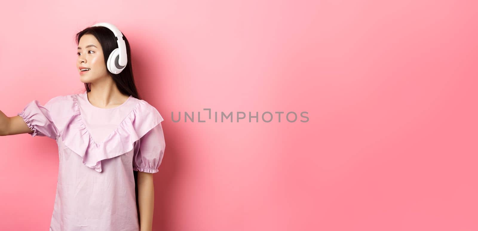 Beautiful asian girl blogger taking selfie in wireless headphones, make photo for social media on smartphone, standing in dress against pink background.