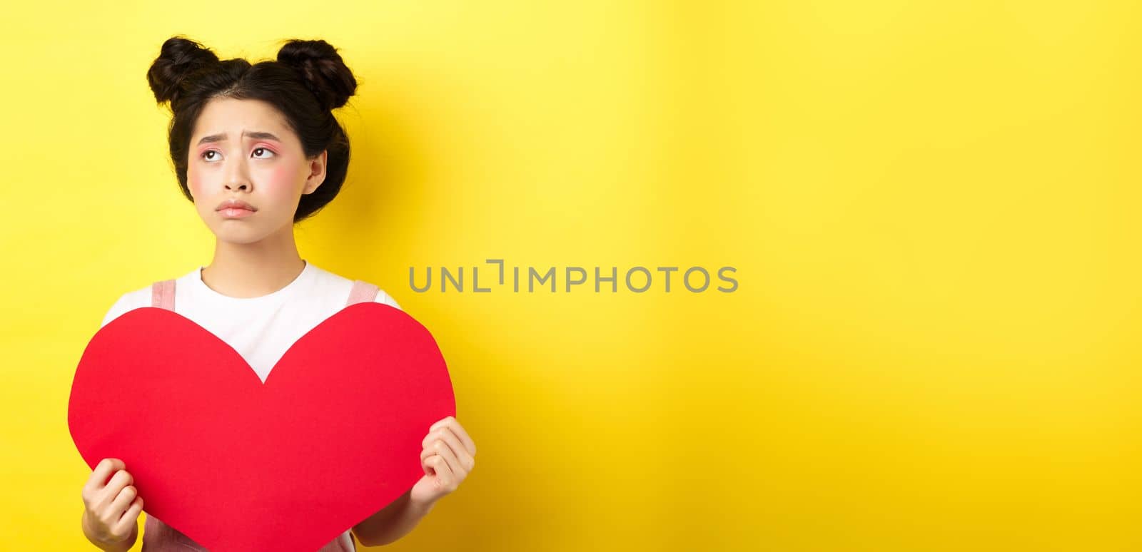 Sad and lonely asian girl standing in make-up with big red heart cutout, left alone on Valentines day, looking left disappointed or upset, standing on yellow background.