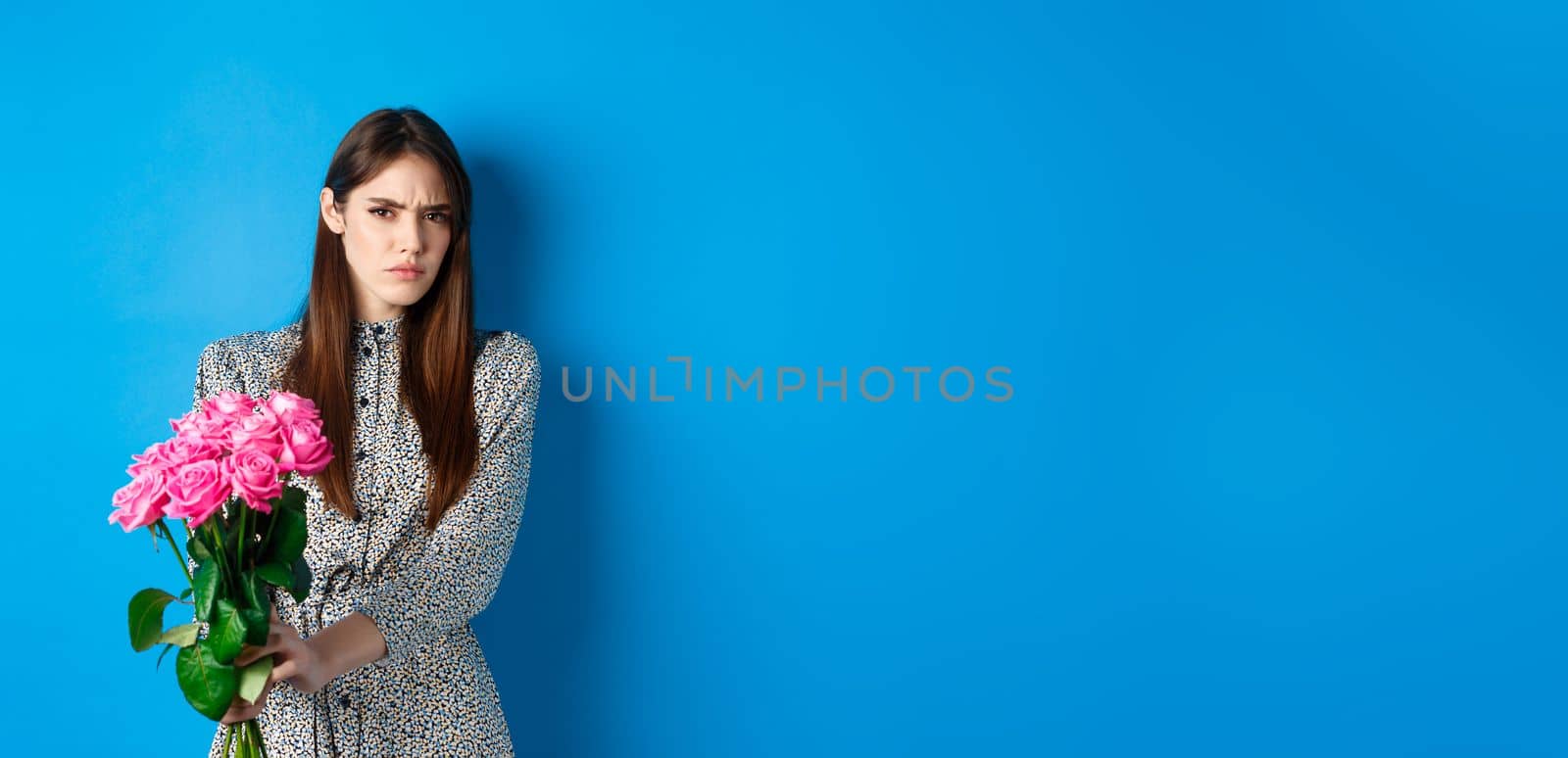 Valentines day concept. Displeased young woman rejecting flowers gift, frowning and grimacing, refusing take roses, standing on blue background.