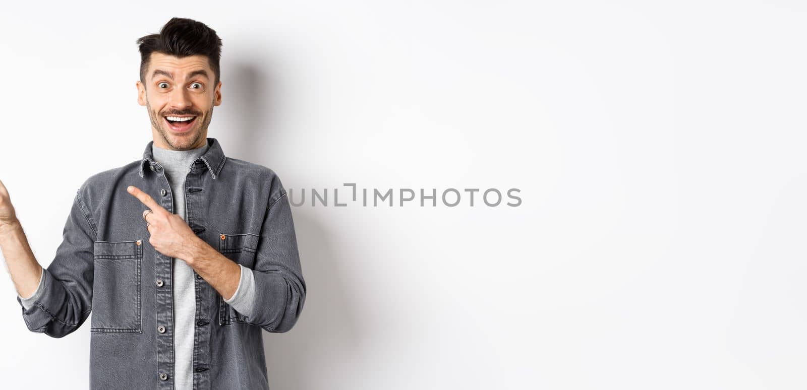 Excited guy with moustache pointing fingers left, showing advertisement and smiling happy, standing on white background.