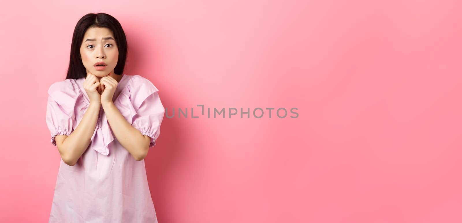 Scared timid girl looking worried at camera, standing concerned on pink background.