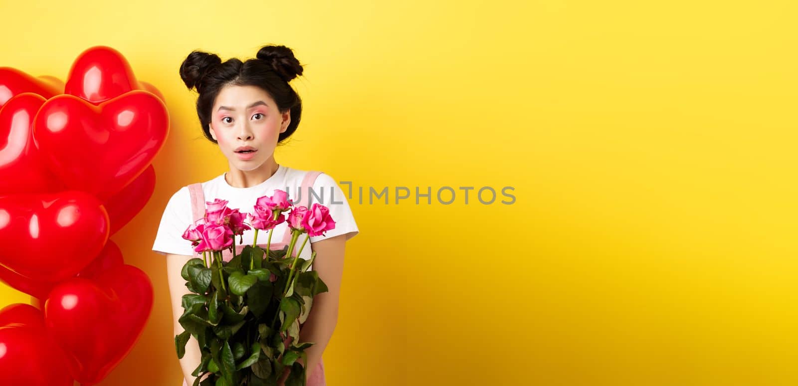Happy Valentines day. Surprised asian girl receive bouquet of pink roses from lover, looking with awe at camera, standing near red heart balloons, yellow background by Benzoix