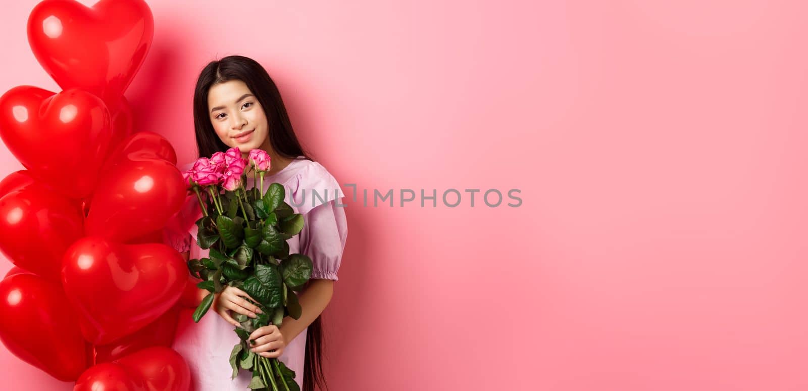 Valentines day concept. Tender and gentle teenage girl holding romantic flowers, standing near red heart balloons from lover and gazing with love, standing on pink background.