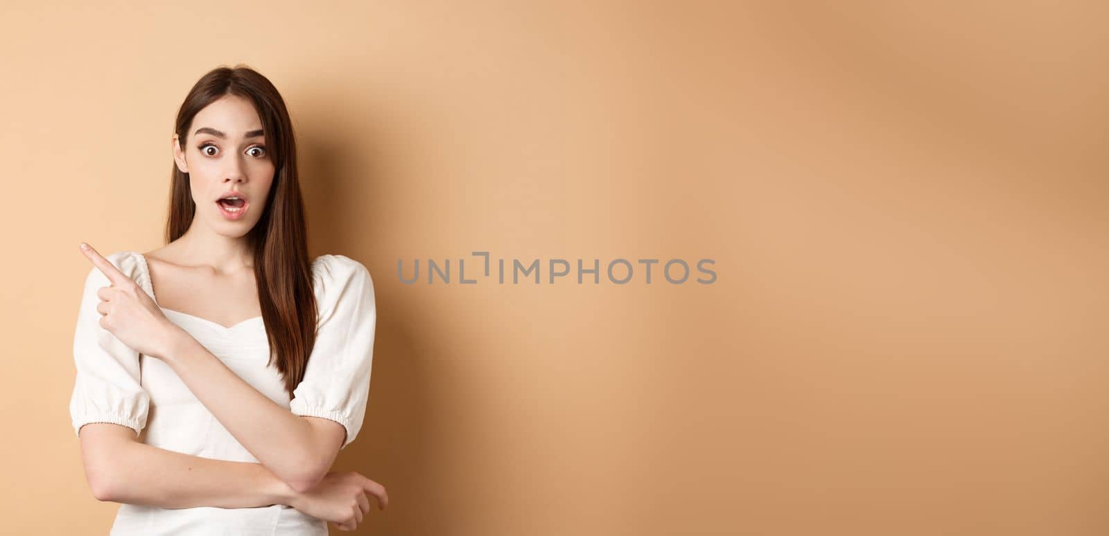 Surprised young girl say wow, pointing finger left at interesting banner, standing curious against beige background.