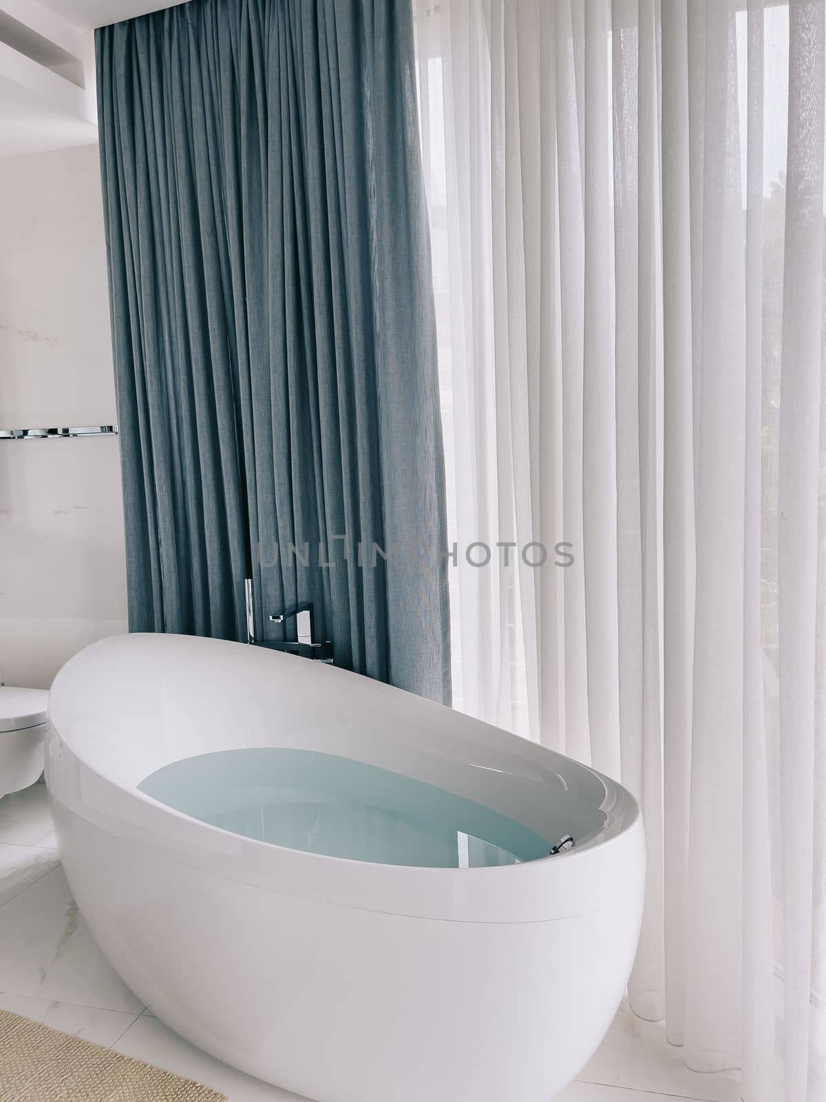 Oval bathtub with chrome taps stands near a panoramic window with curtains. High quality photo
