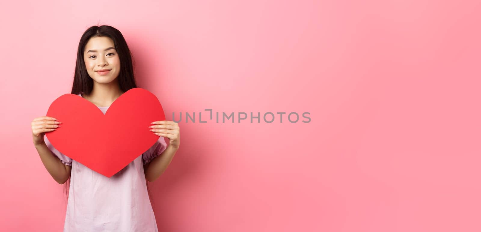 Valentines day concept. Cute teenage asian girl showing big red heart card, falling in love, going on romantic date in dress, smiling tender at camera, pink background by Benzoix