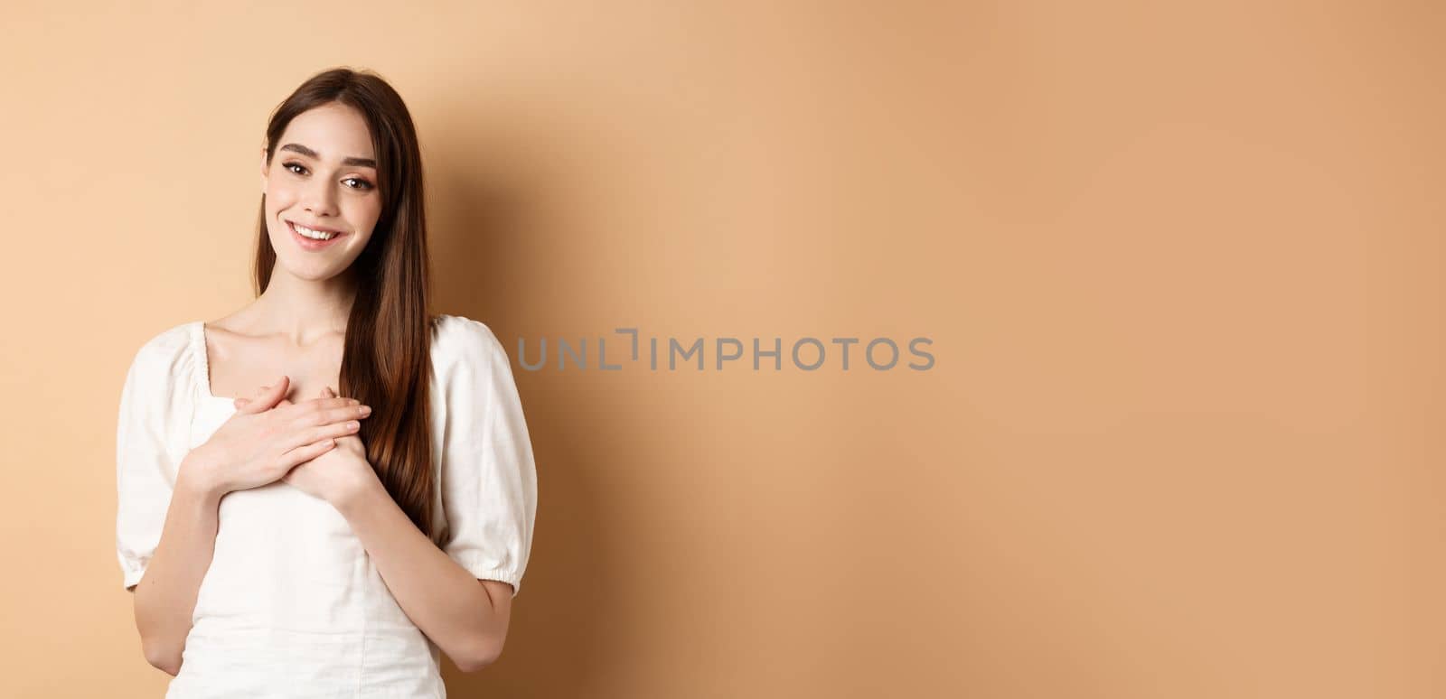 Pretty girl holding hands on heart and smiling, thanking you, feeling grateful and pleased, standing in dress on beige background.