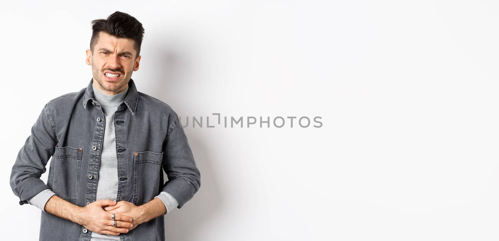 Man with stomach ache frowning and touching belly, having food poison, suffer diarrhea, standing on white background.