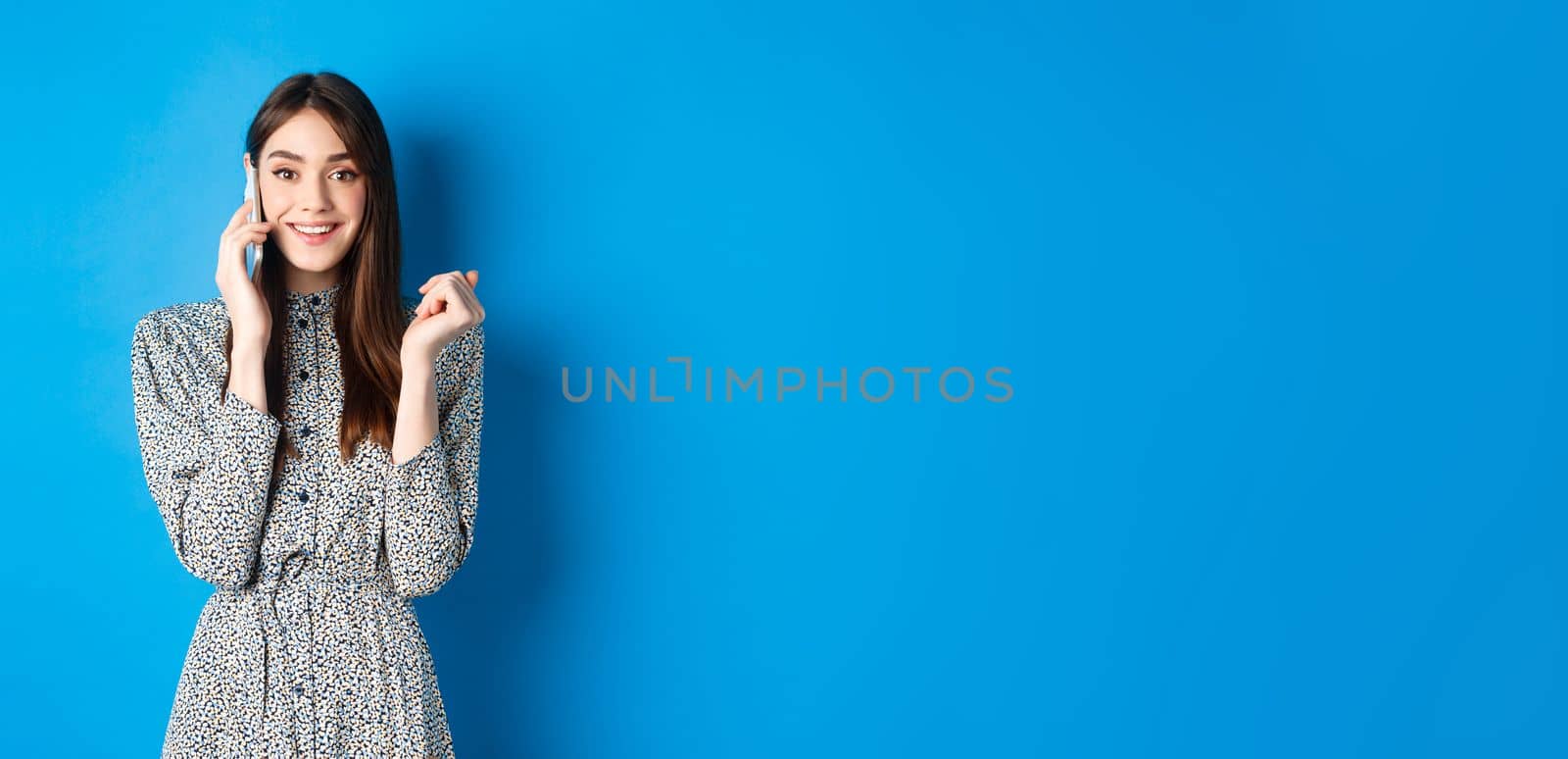 Excited woman receive good news on mobile phone, smiling happy, talking on smartphone, standing on blue background.