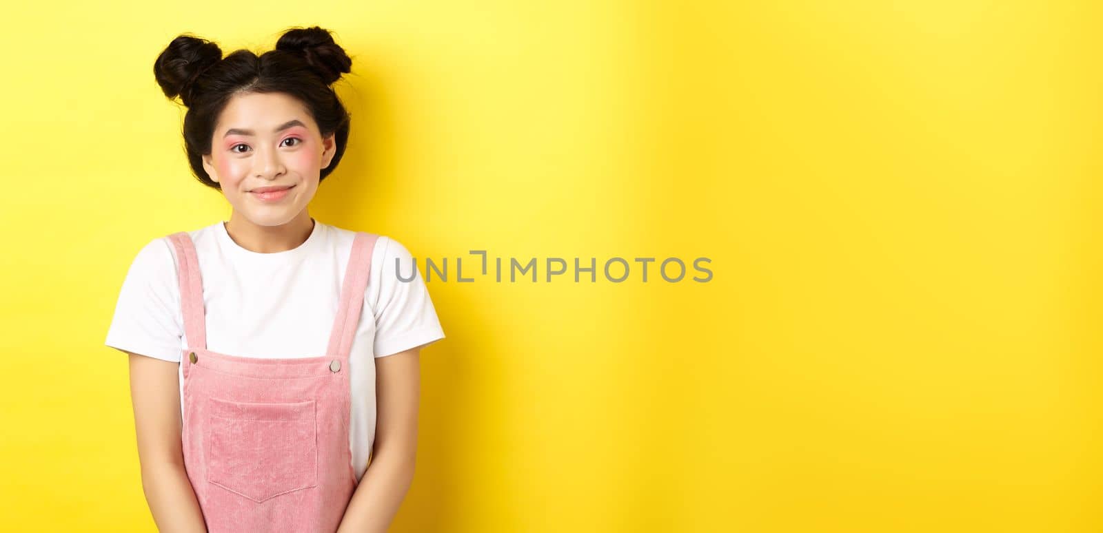Cute asian woman with makeup and summer clothes, smiling silly and happy at camera, yellow background.