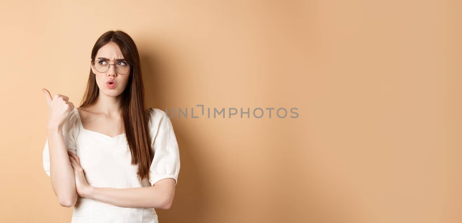 Confused and suspicious young woman in glasses frowning, pointing and looking aside with displeased face expression, standing on beige background.