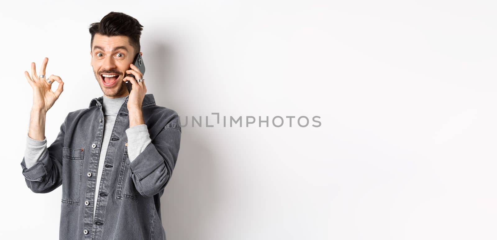 Excited young man talking on phone and showing okay gesture, smiling satisfied, standing against white background.