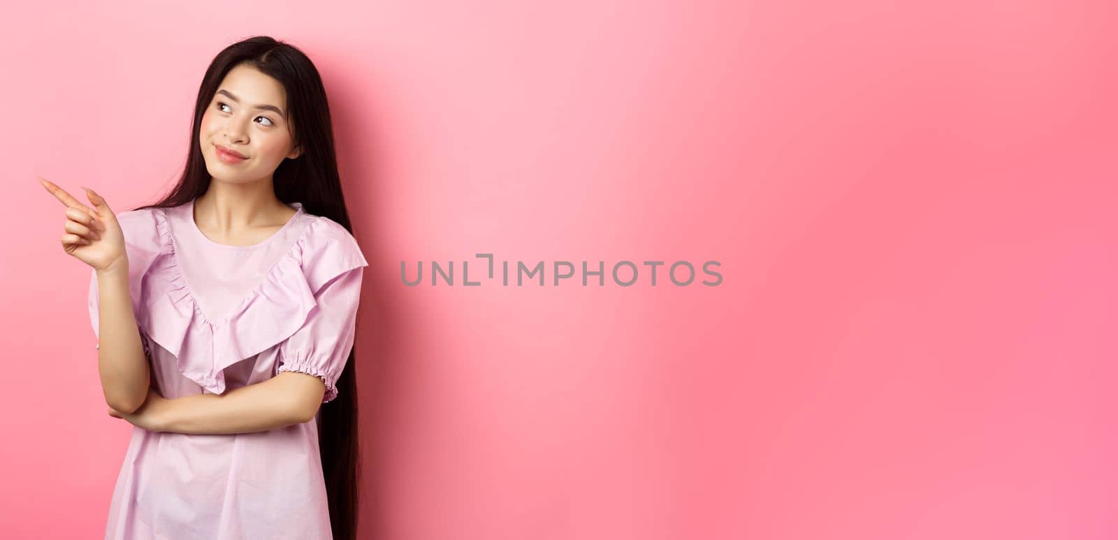 Dreamy asian girl in dress, looking and pointing left at logo, gazing at planner, standing against pink background by Benzoix