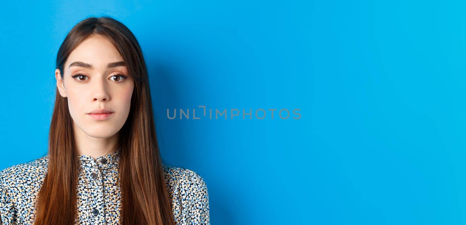 Close-up of candid beautiful woman with long natural hair and makeup, looking at camera, standing on blue background.