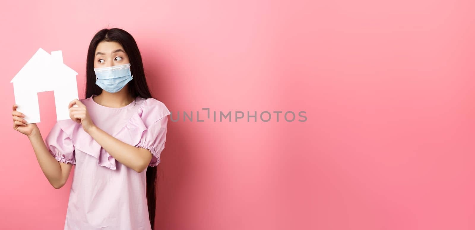 Real estate and pandemic concept. Cute asian woman in medical mask showing paper house cutout, searching for property. standing against pink background.