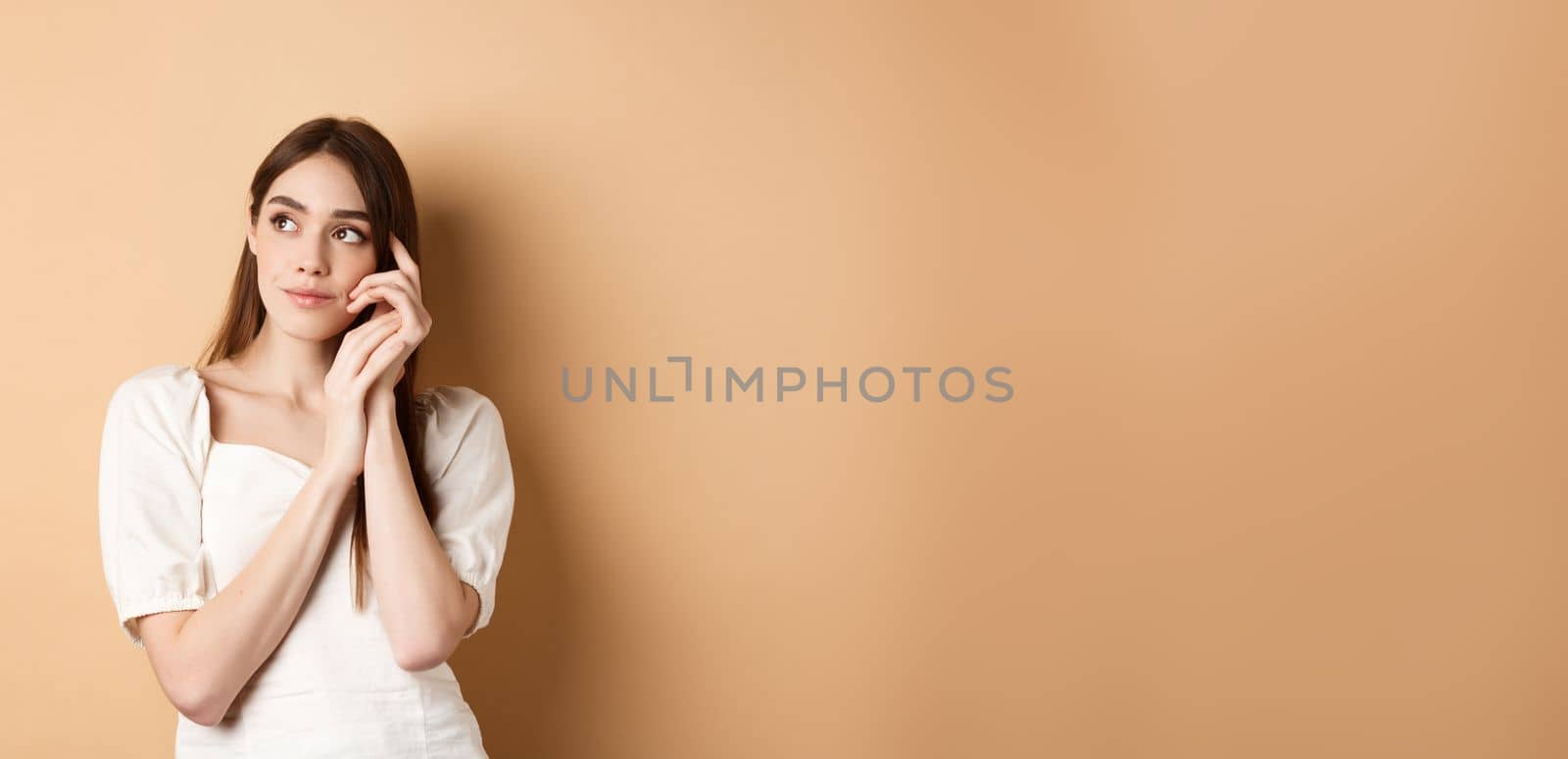Dreamy and romantic girl looking aside with thoughtful gaze, touching clean gentle facial skin, standing against beige background.