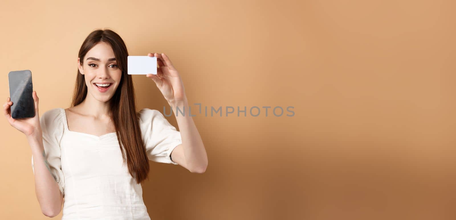 Cheerful young woman showing plastic credit card and empty mobile phone screen, smiling pleased at camera, standing on beige background.