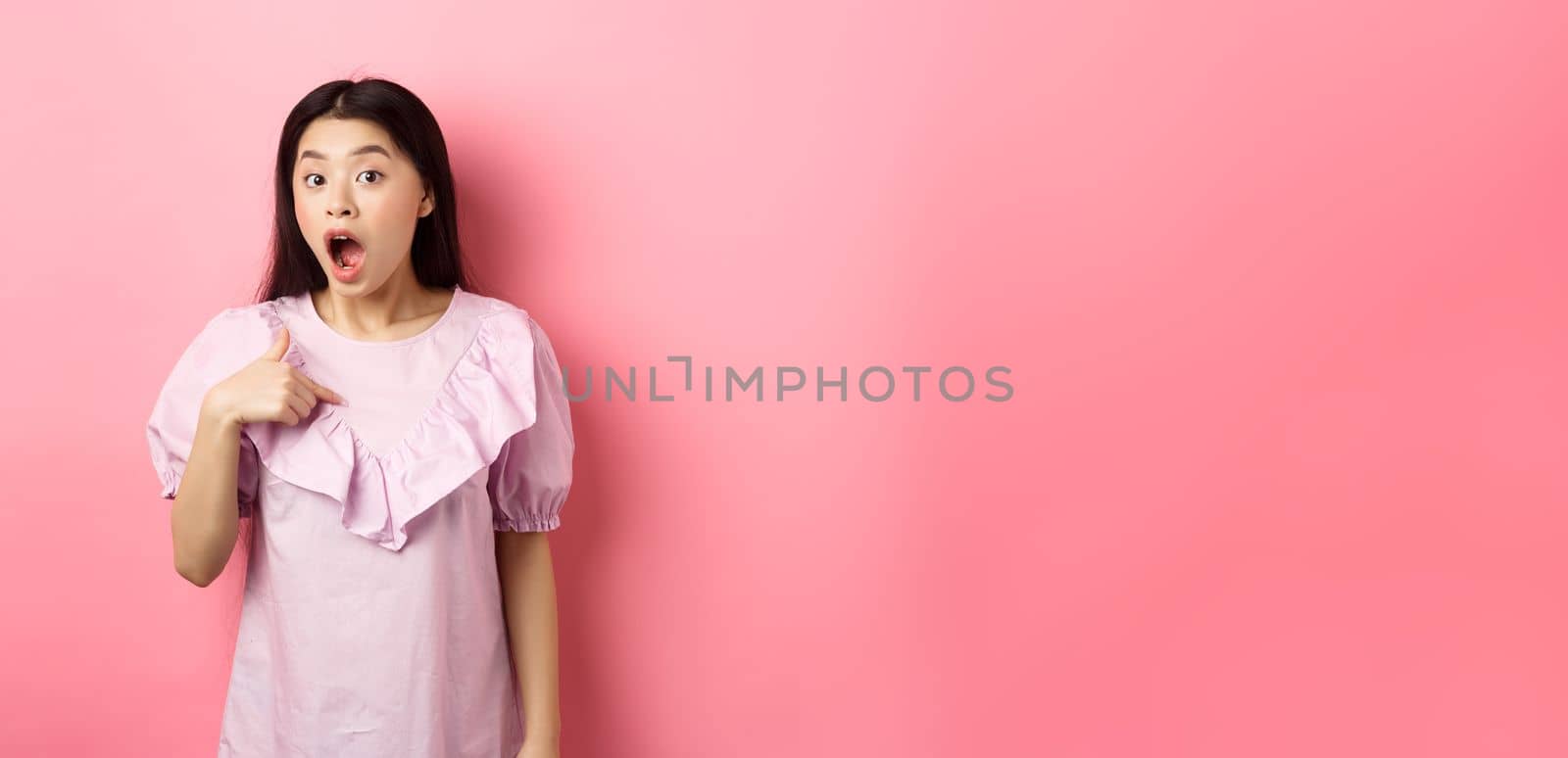 Surprised asian girl gasping wondered, pointing at herself, being chosen or picked, standing in dress against pink background.