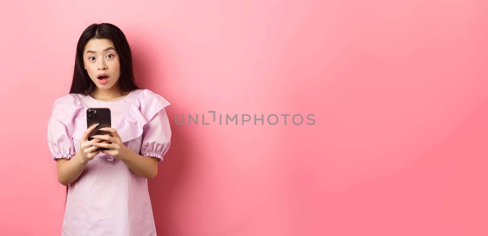 Online shopping. Surprised asian woman in cute dress, open mouth amazed after reading news on smartphone, standing on pink background by Benzoix
