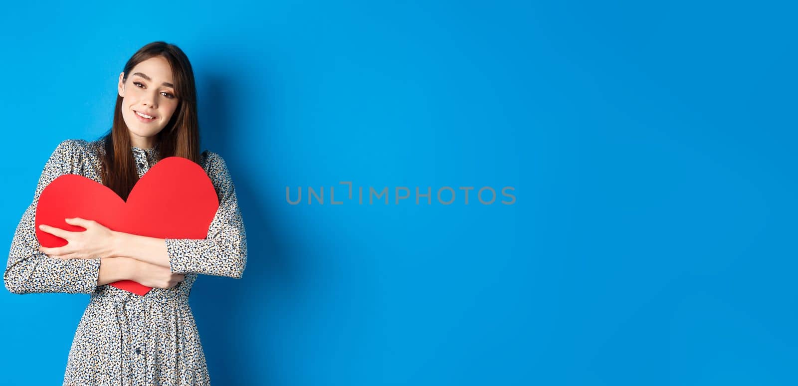 Valentines day. Dreamy romantic woman hugging big red heart cutout, looking sensual at camera, standing in dress on blue background by Benzoix