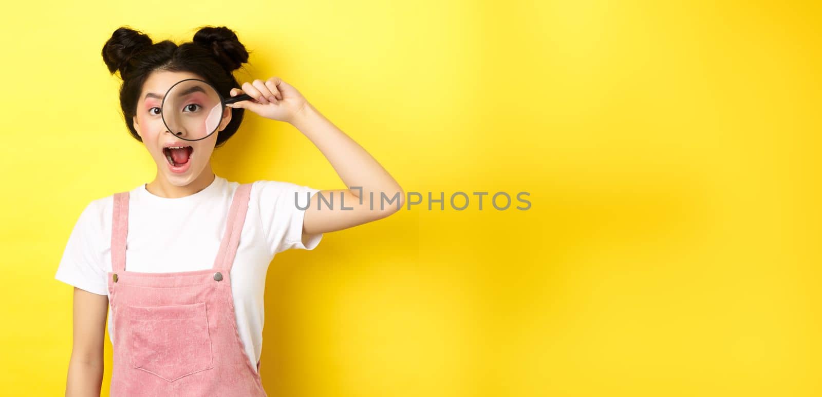 Young asian girl with bright makeup look through magnifying glass, smiling excited, seeing interesting promo, yellow background.