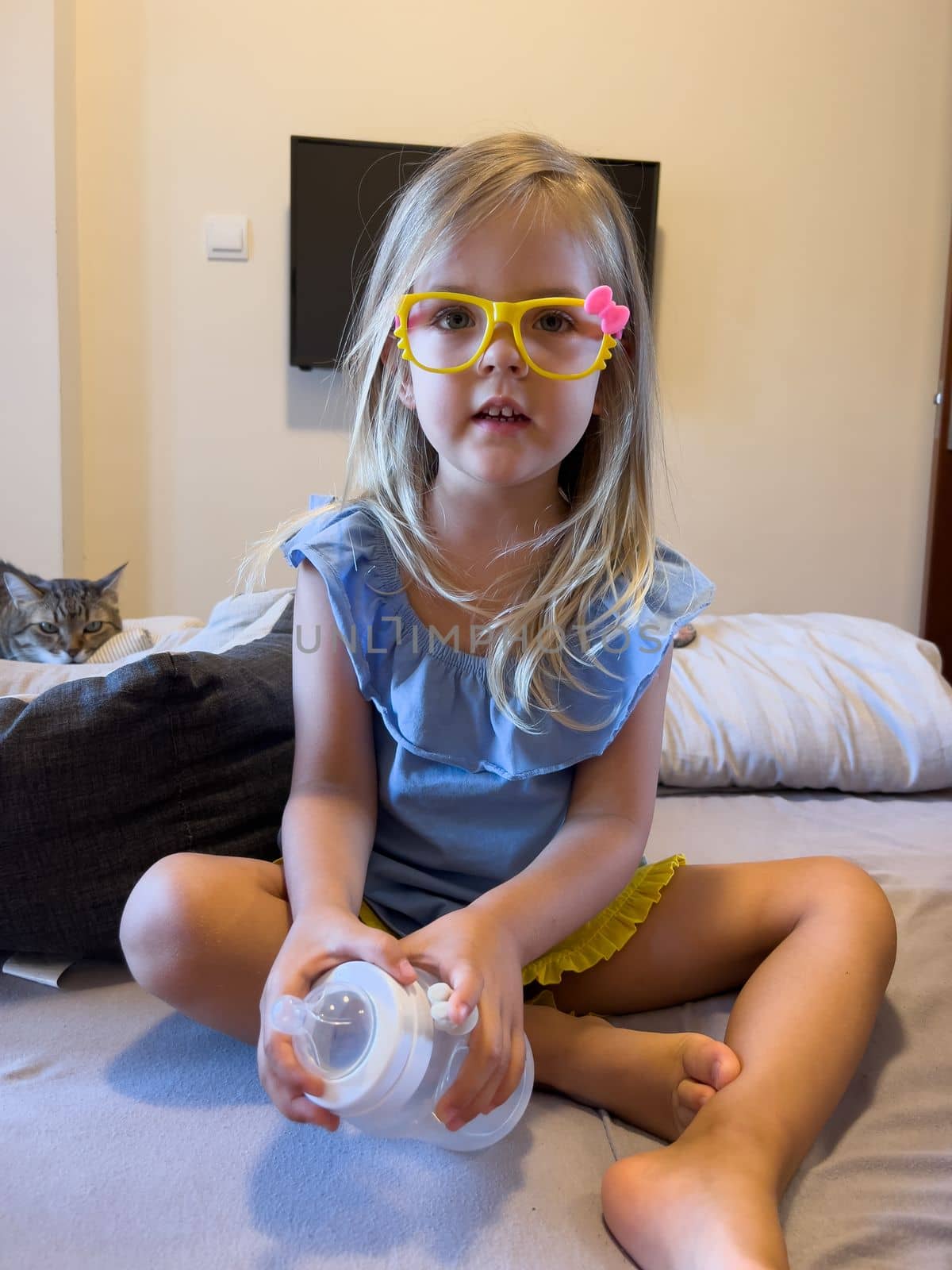 Little girl in glasses with a bottle with a pacifier sits on the bed. High quality photo
