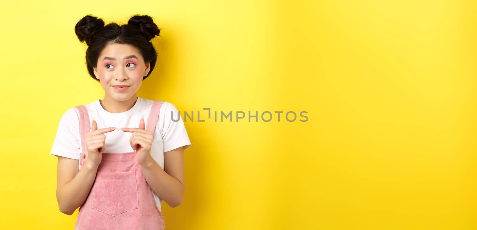 Summer lifestyle concept. Cute shy asian girl looking away and smiling silly, avoiding eye contact, standing against yellow background.