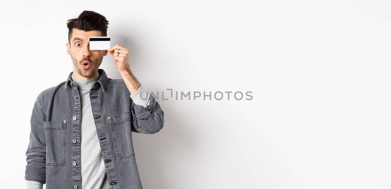 Excited funny man hold plastic credit card on eye and say wow, checking out special deal, standing on white background.