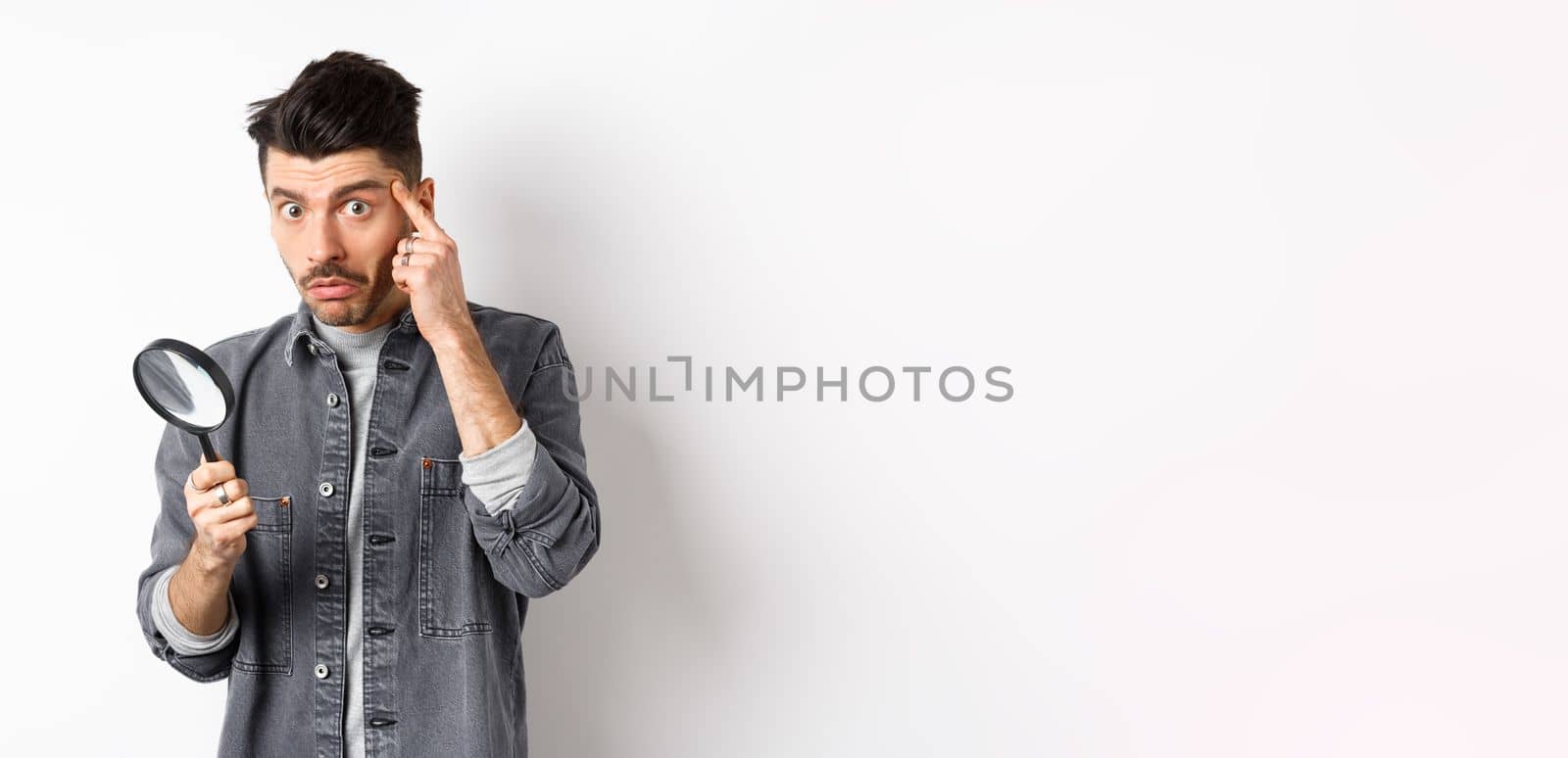Confused guy found something down with magnifying glass, look puzzled at camera, standing against white background.