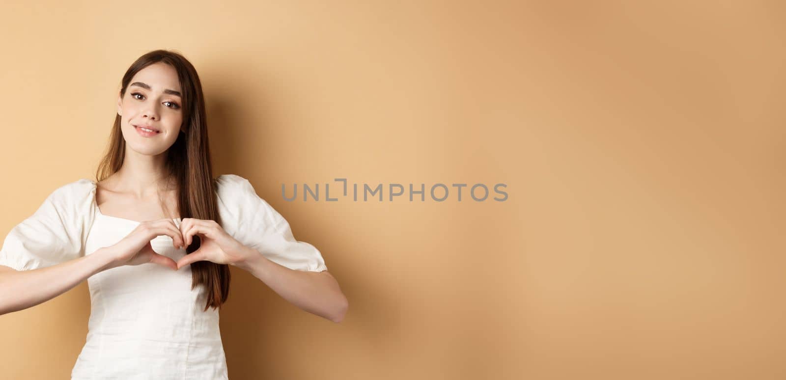 Beautiful caucasian woman say I love you, showing heart gesture and smile at camera, beige background. Concept of romance and Valentines day.