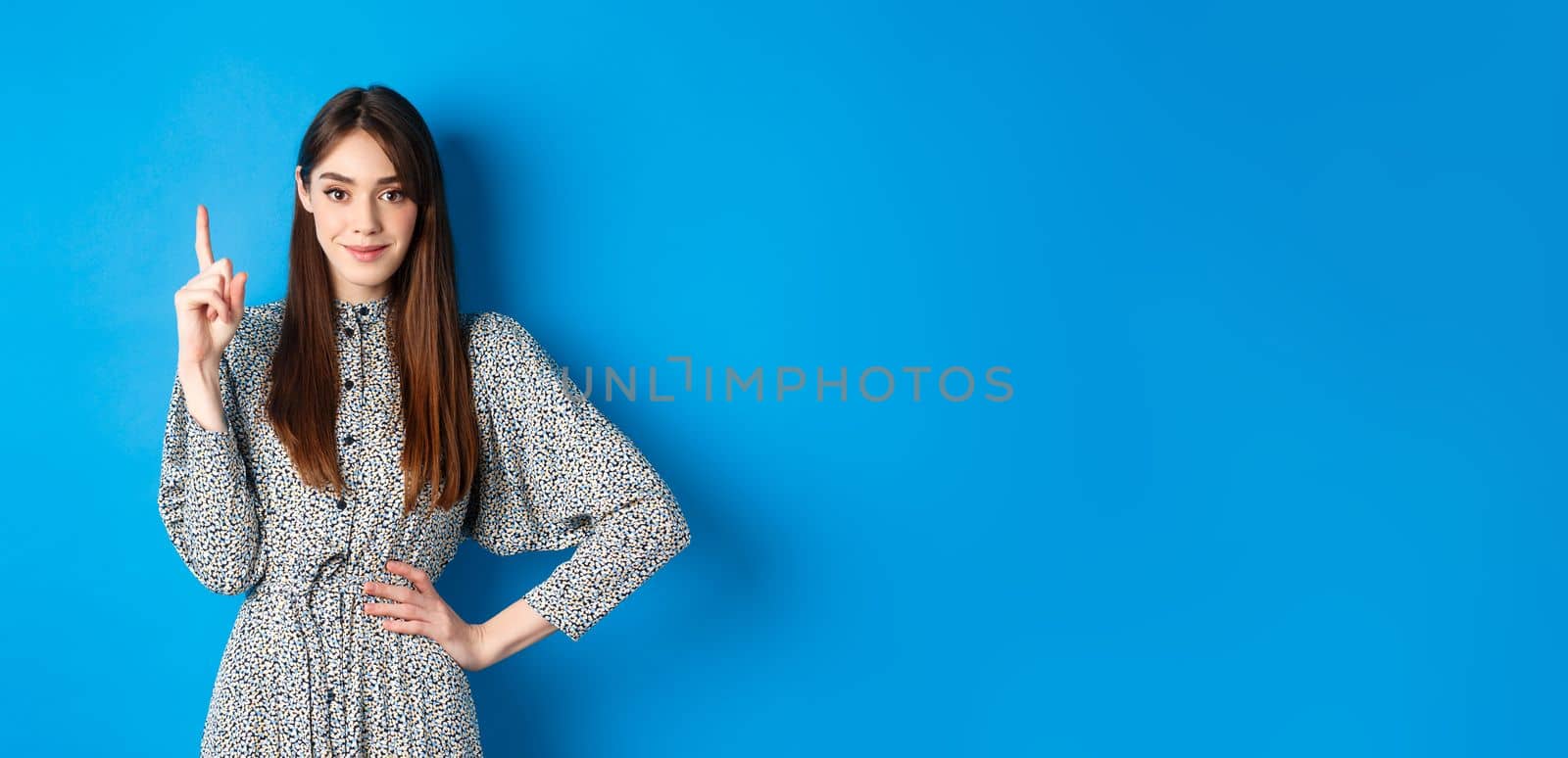 Young beautiful lady in dress with natural long hair, showing number one with finger and smiling, standing against blue background.