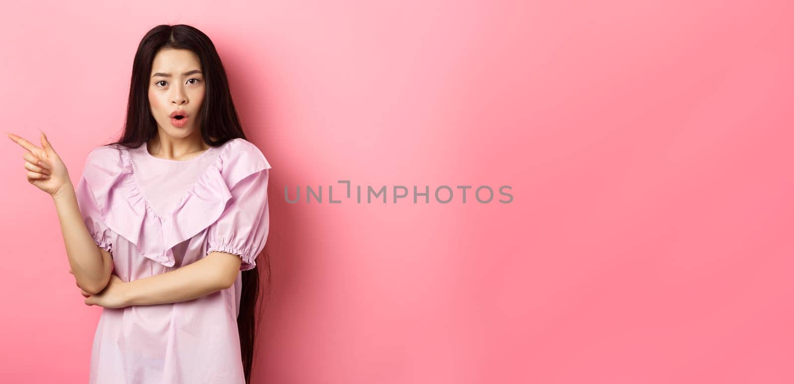 Shocked asian teen girl in dress pointing finger left, gasping wondered and look curious at camera, asking question about promo, standing on pink background by Benzoix