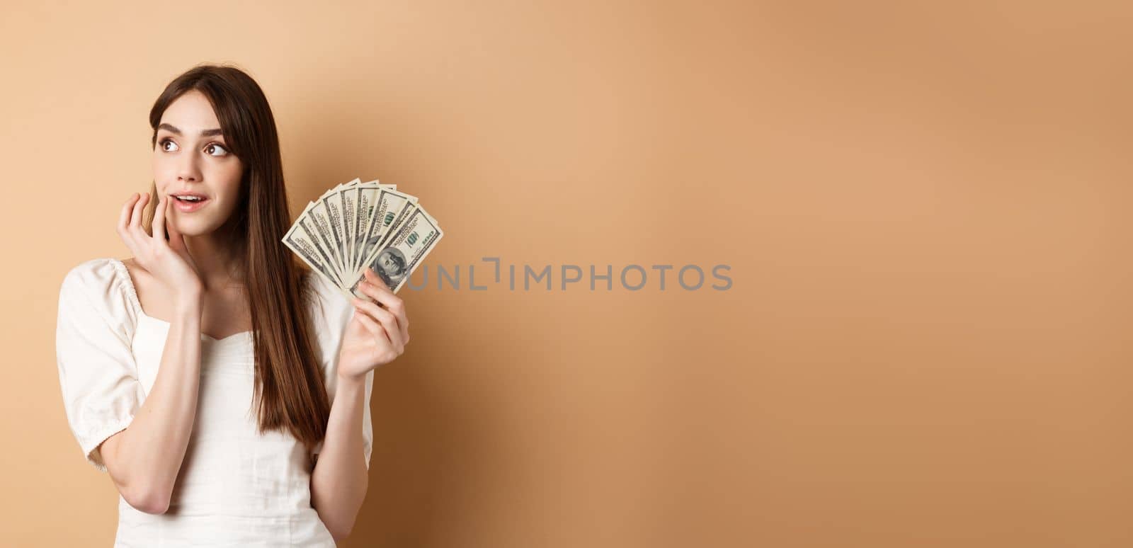 Dreamy girl holding dollar bills and thinking off shopping, looking at upper left corner thoughtful, plan how to waste money, standing on beige background.