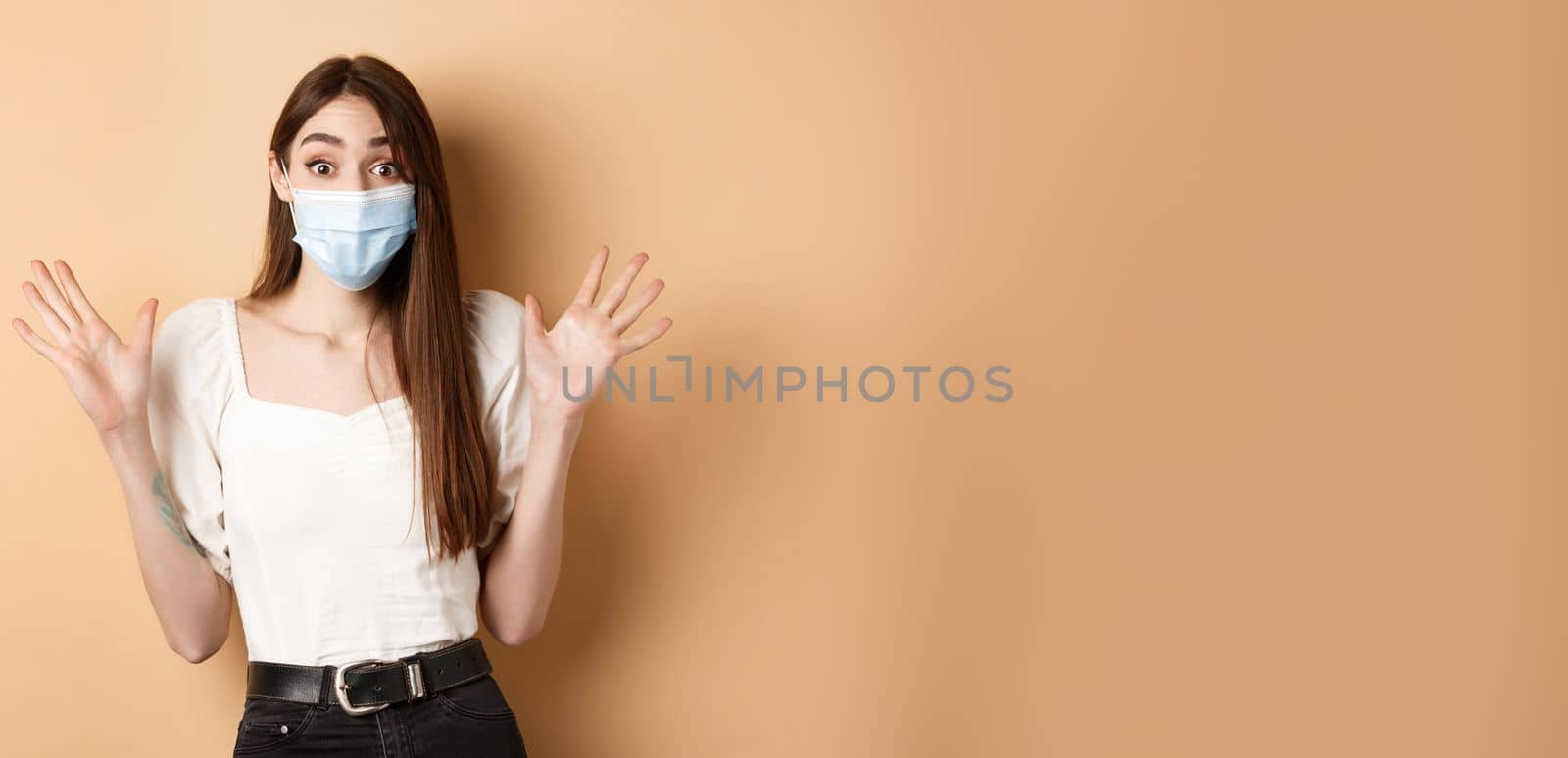 Covid-19 and lifestyle concept. Surprised woman in medical mask scream excited, raising hands up and shouting big news, standing on beige background.