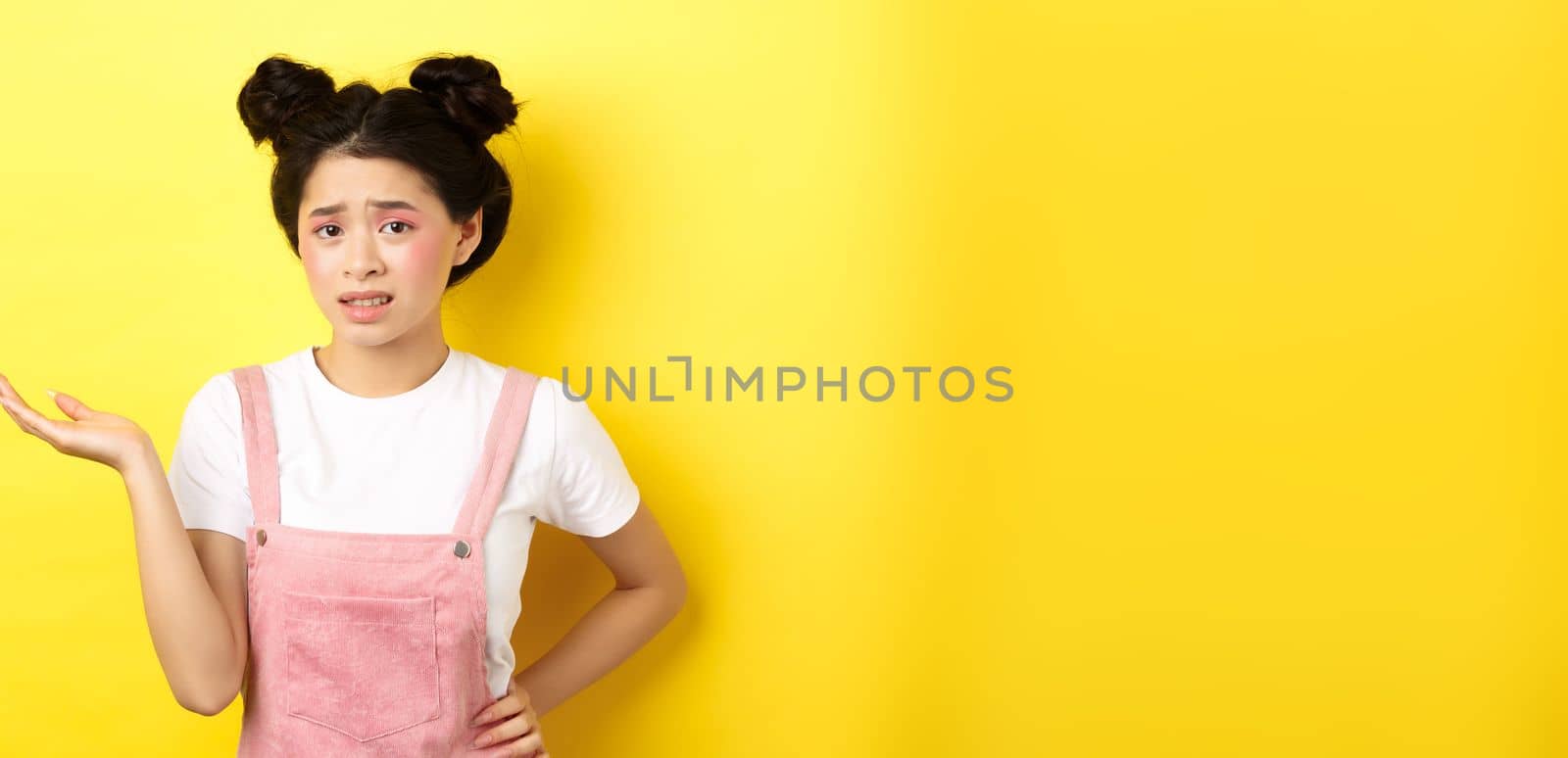 Confused teen asian girl with bright makeup and summer clothes, raising hand up and frowning puzzled, cant understand, yellow background.