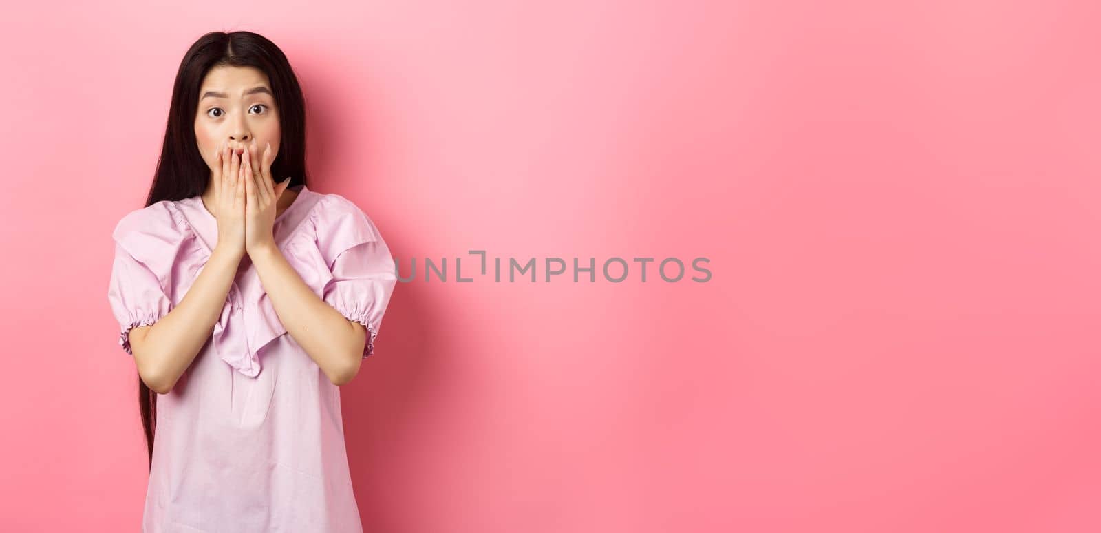 Shocked asian girl with long dark hair, gasping and covering mouth with hands, look at bad news, terrible accident, standing on pink background by Benzoix