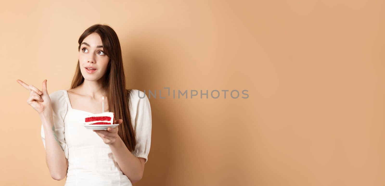 Thoughtful birthday girl holding cake with candle, thinking of wish and pointing left at logo, standing on beige background.