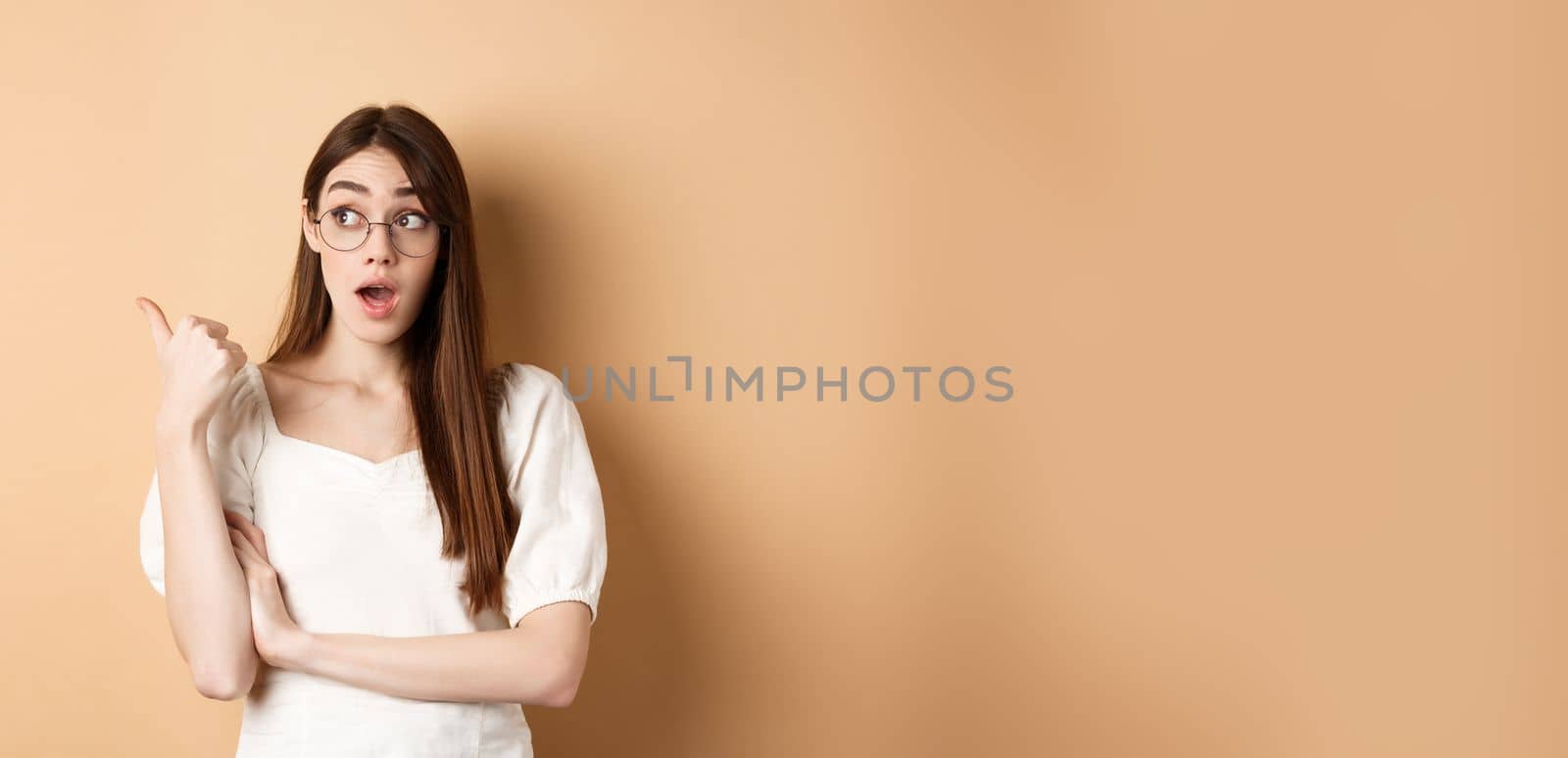 Excited girl in glasses pointing left, looking surprised at banner, standing on beige background.