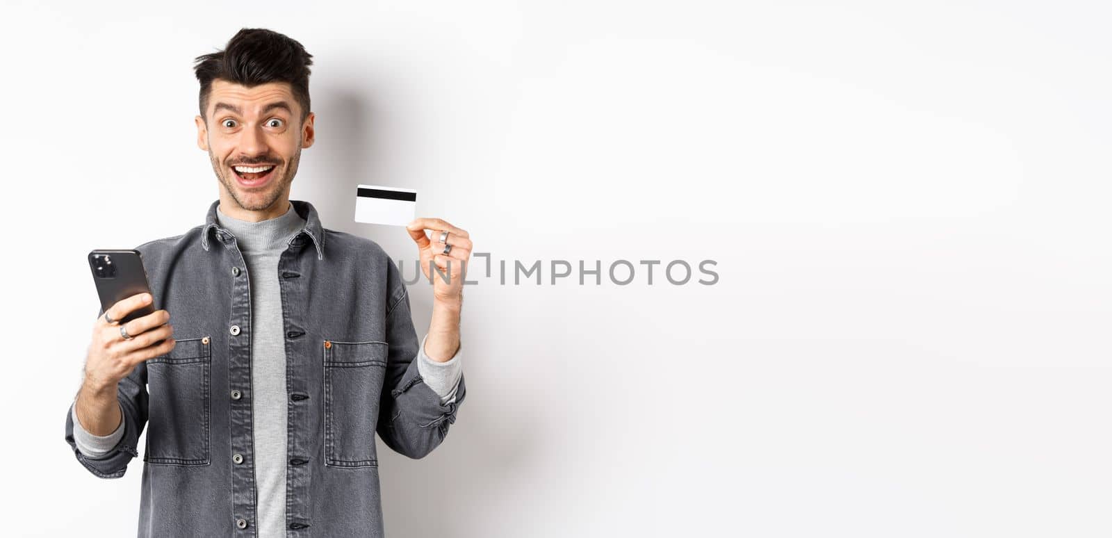 Online shopping concept. Excited man making purchase in internet, easy buying with plastic credit card and smartphone, smiling happy at camera, white background.