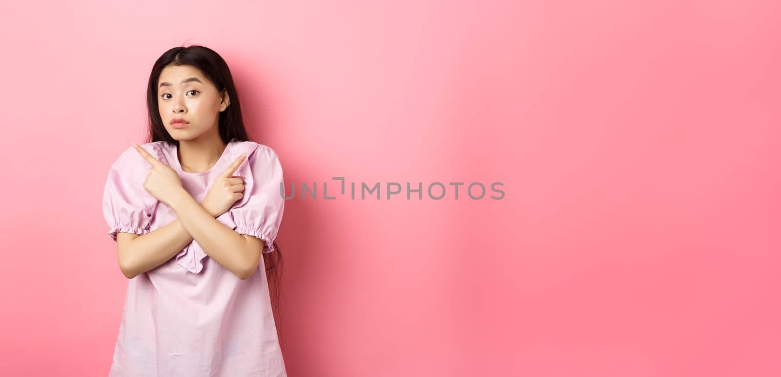 Clueless cute asian girl pointing sideways and look confused, cant make decision, standing hesitant against pink background by Benzoix