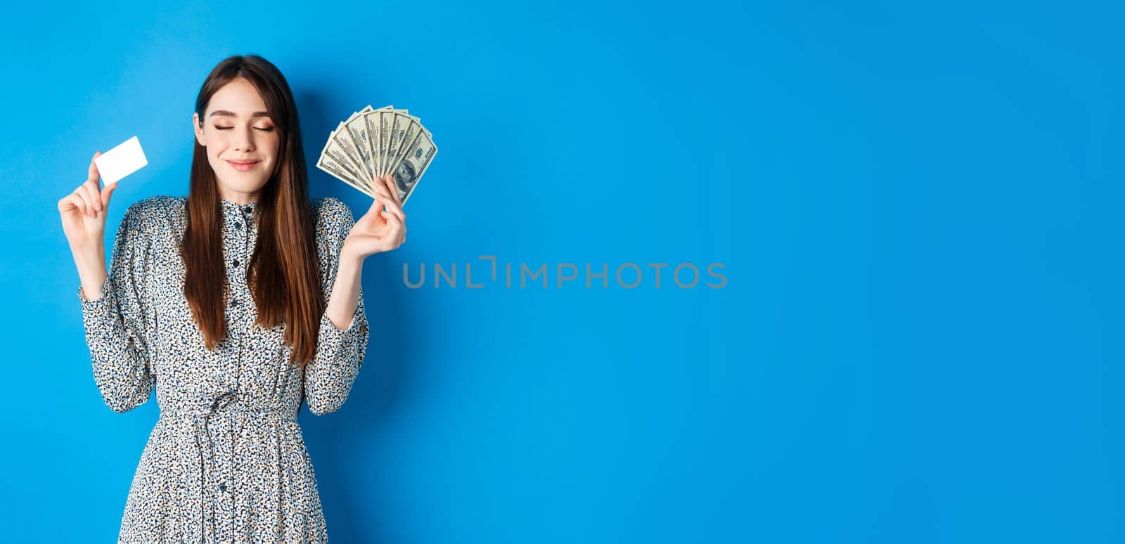 Shopping. Happy smiling woman looking satisfied with eyes closed, showing money dollar bills and plastic credit card, standing dreamy against blue background by Benzoix