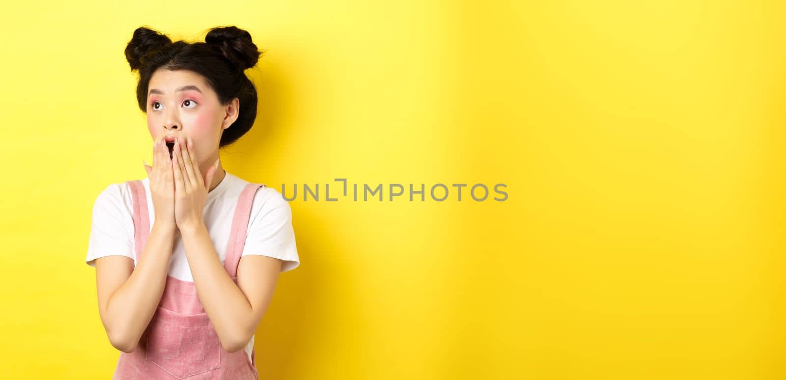 Shocked teen asian girl with makeup, gasping and covering mouth, looking left at logo amazed, standing on yellow background.